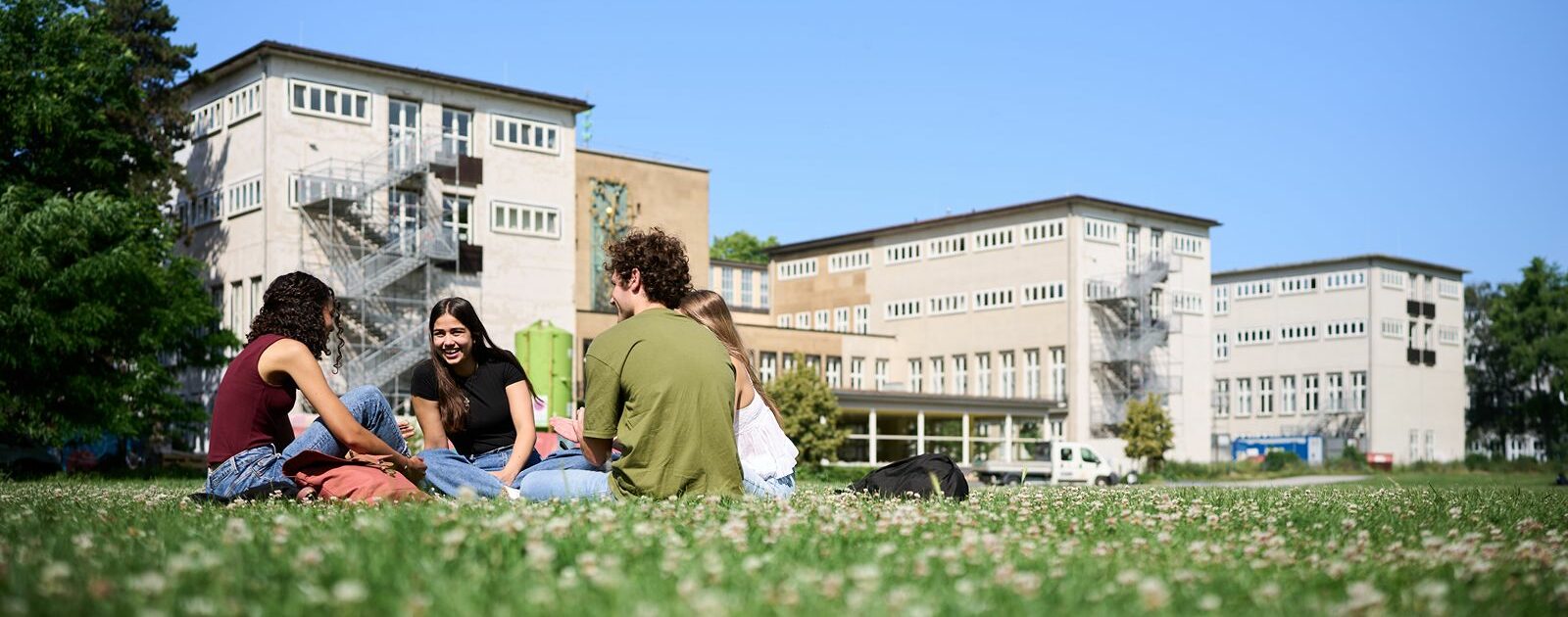 Eine Gruppe von Studierenden sitzt auf einer Wiese vor der Universit&auml;t und unterh&auml;lt sich