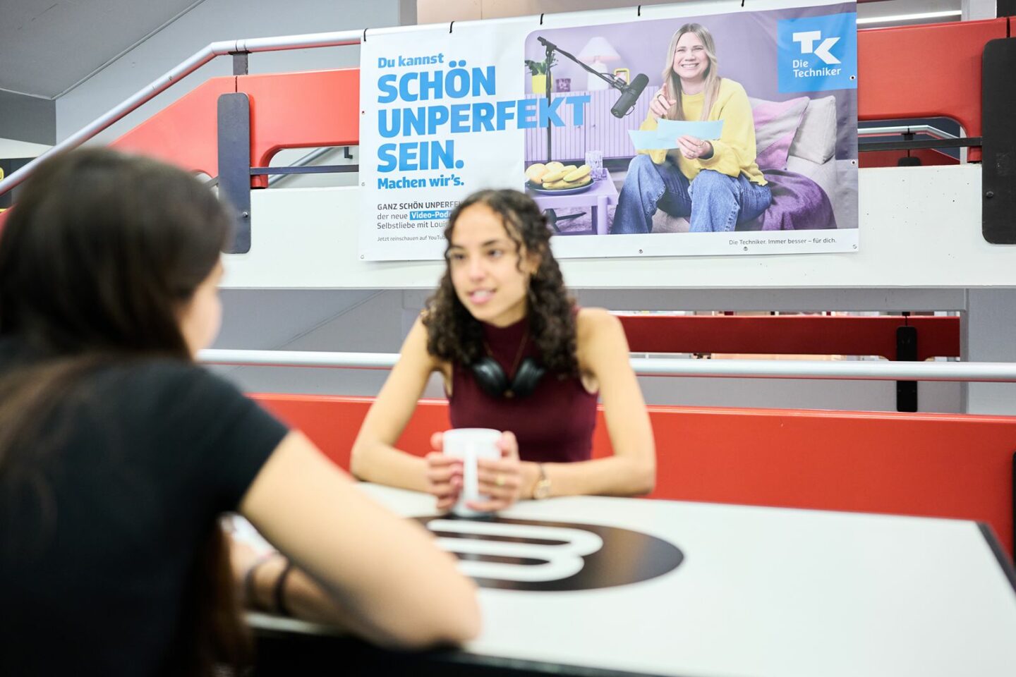 A tensioned banner hangs in the stairwell; in front of it, two female students are sitting and talking.